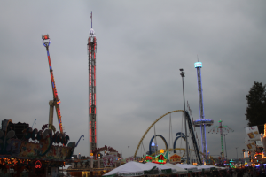 Eine Menschenmenge versammelt sich um einen nächtlichen Fasching, beleuchtet von bunten Lichtern von Fahrgeschäften, Pfählen und Bäumen, unter einem bewölkten Himmel.