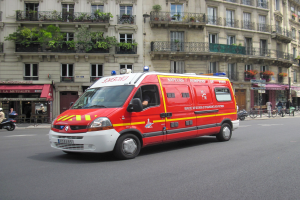 Ein Rettungswagen steht auf der Straäenseite vor einem Geb"ude mit Fenstern, Gel"andern und Pflanzen, mit einigen Menschen und Fahrr"adern im Hintergrund.