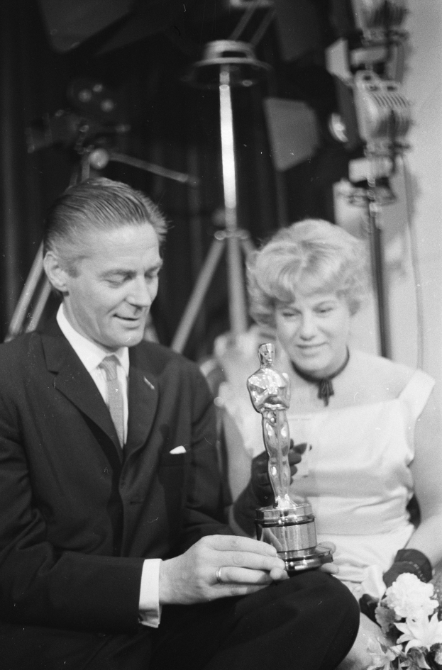 Ein Mann in einem schwarzen Blazer, weißem Hemd und Krawatte hält eine Oscar-Statue in der Hand und sitzt neben einer Frau, die einen Blumenstrauß hält, beide in festlicher Kleidung, mit Lichtern im Hintergrund, in Schwarz-Weiß.