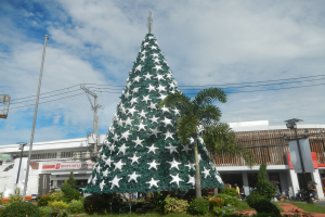 Ein großer Weihnachtsbaum steht in der Mitte einer Stadtstraße, umgeben von Pflanzen, Bäumen, Pfählen, Drähten und Gebäuden, mit ein paar Menschen und Fahrzeugen im Hintergrund unter einem bewölkten Himmel.