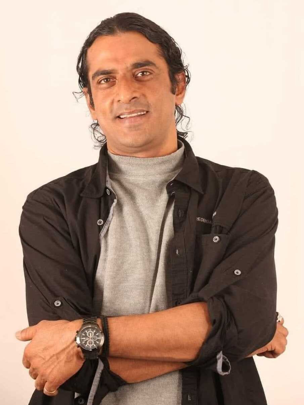 Ein Mann in einem schwarzen und aschfarbenen Kleid mit überschlagenen Armen, der eine Uhr tr├Ągt und gegen einen wei├čen Hintergrund steht, der in Gedanken versunken zu sein scheint.