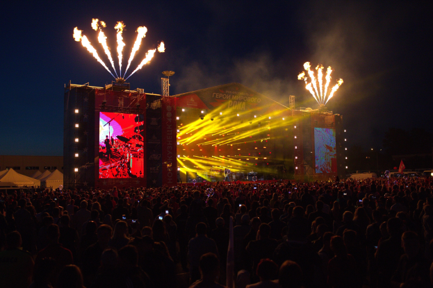 Eine große Menschenmenge auf einem Musikfestival schaut Zuschauern auf einer hell erleuchteten Bühne zu, mit Zelten links und Feuerwerk am dunklen Himmel dahinter.