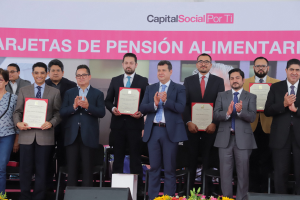 Eine Gruppe von Menschen auf einer Bühne, die klatschen und Zertifikate halten, mit Stühlen in einem Halbkreis darum herum und einem Banner mit der Aufschrift "Capital Social for Ti" im Hintergrund, zusammen mit Blumenbouquets und Rednern unten.