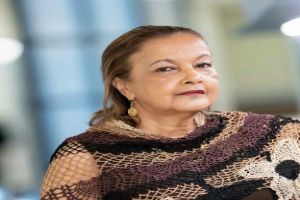 Eine Frau in einem schwarzen Kleid und einer braunen Schälle steht an einem Fenster und sieht nachdenklich aus, mit einem unscharfen Hintergrund.