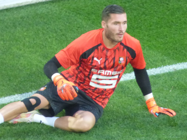 Ein Fußballspieler in rotem T-Shirt, schwarzen Shorts, weißen Socken und orangen Baseball-Handschuhen sitzt auf dem Boden mit den Händen auf den Knien.