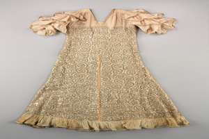 Ein cremefarbenes Kleid mit goldener Stickerei und Rüschen an den Ärmeln, das auf einer weißen Oberfläche präsentiert wird und mit detaillierten Mustern und Ornamenten verziert ist.