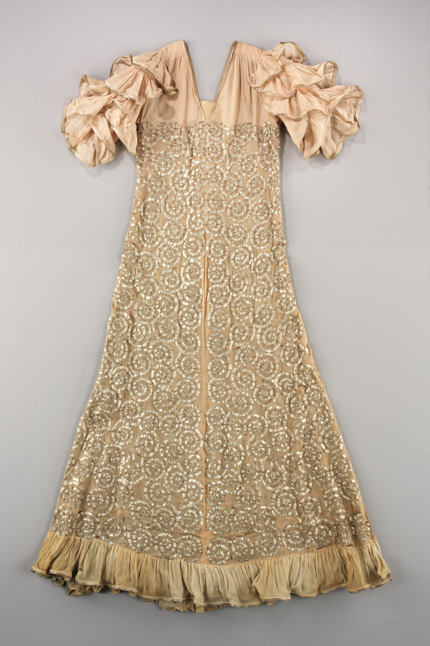 Ein cremefarbenes Kleid mit goldener Stickerei und Rüschen an den Ärmeln, das auf einer weißen Oberfläche präsentiert wird und mit detaillierten Mustern und Ornamenten verziert ist.