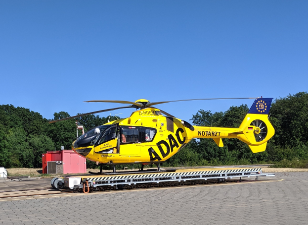 Ein gelber Hubschrauber steht auf einem Flughafen-Asphalt, umgeben von Bäumen und einem klaren blauen Himmel, mit verschiedenen Gegenständen, die mit dem Hubschrauber in Verbindung stehen, drumherum verstreut.