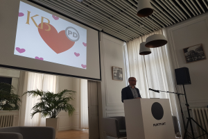 Ein Mann steht an einem Rednerpult mit einem Mikrofon und hält eine Präsentation zum Thema Geschlechtergerechtigkeit, mit einem Projektionsschirm dahinter, einem Lautsprecher, Stühlen, einem Topf mit einer Pflanze, gerahmten Fotos an der Wand, Vorhängen und Deckenleuchten.