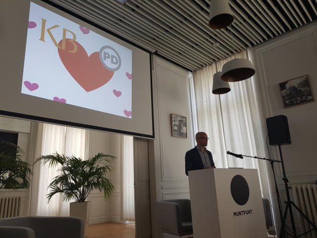 Ein Mann steht an einem Rednerpult mit einem Mikrofon und hält eine Präsentation zum Thema Geschlechtergerechtigkeit, mit einem Projektionsschirm dahinter, einem Lautsprecher, Stühlen, einem Topf mit einer Pflanze, gerahmten Fotos an der Wand, Vorhängen und Deckenleuchten.