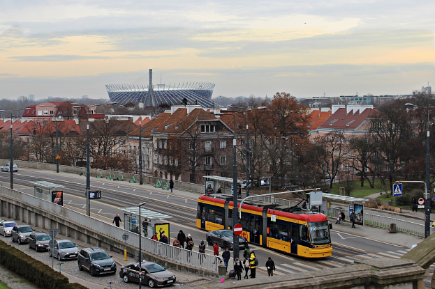 Eine gelbe und schwarze Straßenbahn fährt auf einer von Hochhäusern gesäumten Stadtstraße, umgeben von Autos, Fußgängern, Laternen, Schildern, Bäumen und Gras, mit bewölktem Himmel im Hintergrund.