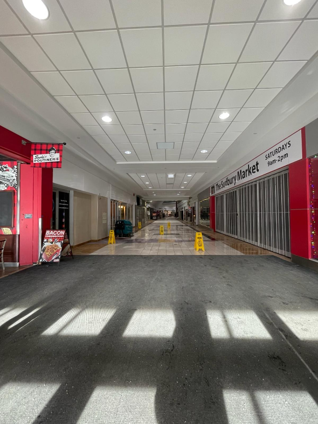 Innenseite eines hell erleuchteten Einkaufszentrums mit einem Stuhl und einer Tafel, die "Sydbury Market - Vorher und Nachher" trägt, auf der linken Seite und verstreuten Gegenständen auf der rechten und auf dem Boden.