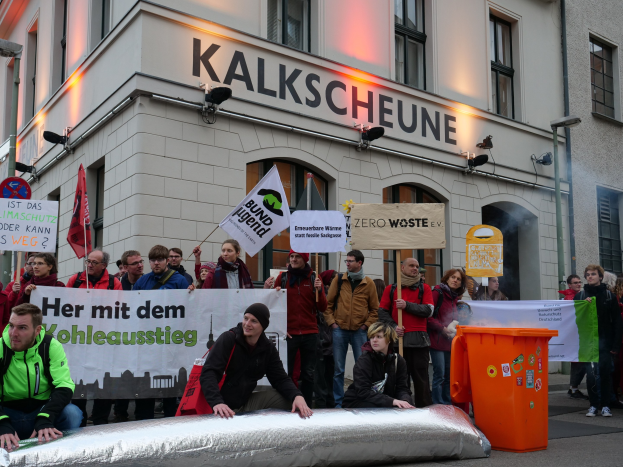 Eine Gruppe von Menschen mit Protestschildern steht vor einem Gebäude mit Fenstern und Schildern, während zwei Personen auf einem Objekt im Vordergrund sitzen und ein Müllcontainer rechts zu sehen ist.