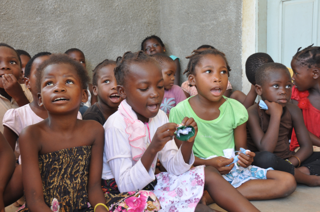 Eine Gruppe von Kindern sitzt auf dem Boden vor einem Gebäude, im Hintergrund eine Wand und eine Tür, ihre Gesichter voller Traurigkeit und Verzweiflung.