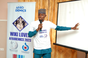 Ein Mann in einer Mütze spricht in ein Mikrofon, während er vor einer Whiteboard steht, mit einem Banner, Vorhang und einer Schalttafel im Hintergrund.