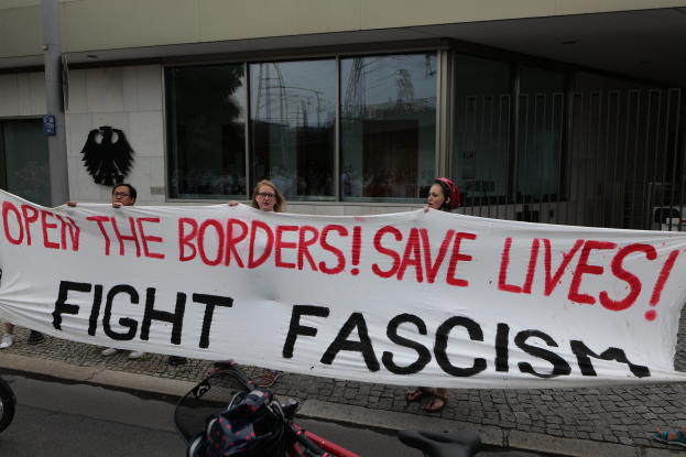 Eine Gruppe von Menschen mit einer Schildern mit der Aufschrift "Öffne die Grenzen, rette Leben, bekämpfe Faschismus" vor einem Gebäude mit Glasfenstern und einem Pfahl, mit einem Fahrrad und einer Tasche auf der Straße.