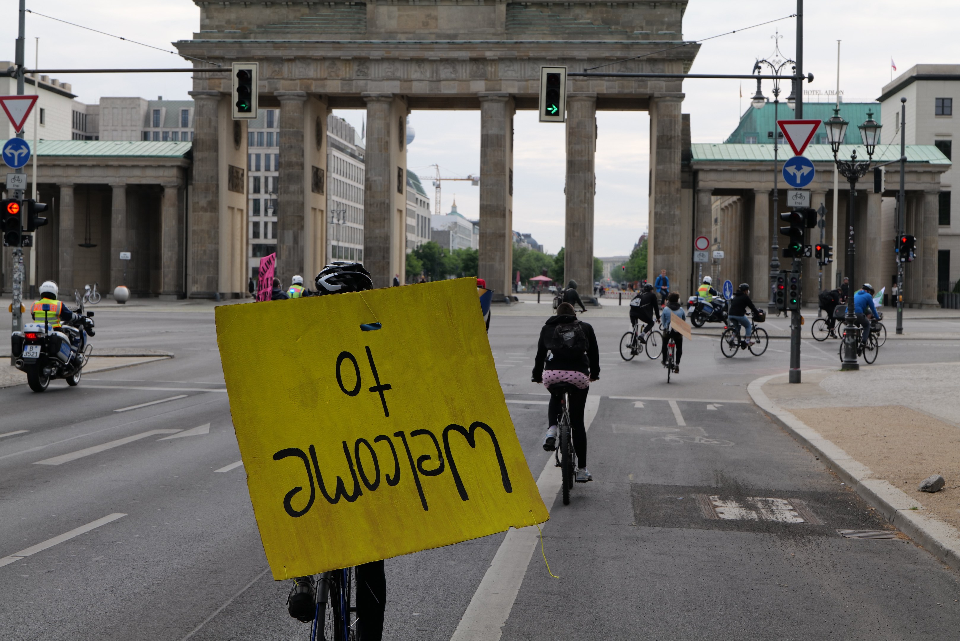 Eine Gruppe von Radfahrern fährt an der Brandenburgertor in Berlin vorbei, einer hält ein gelbes Schild, mit Laternen und Gebäuden im Hintergrund und einem klaren Himmel.