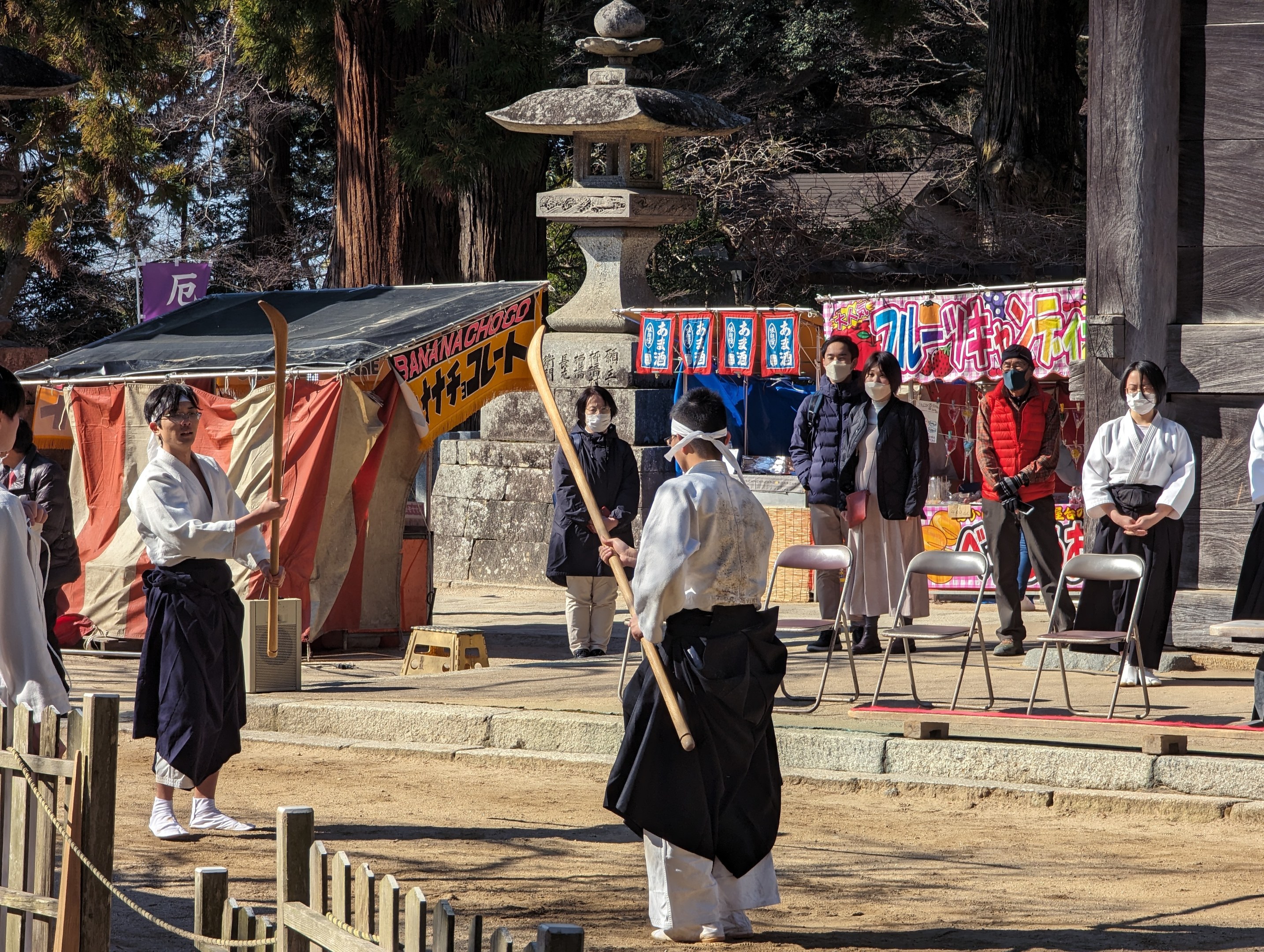 Eine Gruppe von Menschen in formeller Kleidung, einige tragen Masken und halten Stöcke, versammelt sich vor einem Gebäude während einer traditionellen japanischen Zeremonie in Kyoto, mit Stühlen, einem Zelt, Bannern, Bäumen und einem klaren blauen Himmel im Hintergrund.