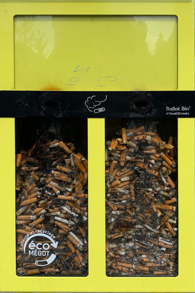 Gelber Müllcontainer voller Zigarettenstummeln auf einem Gehweg, beschriftet für Zigarettenentsorgung.