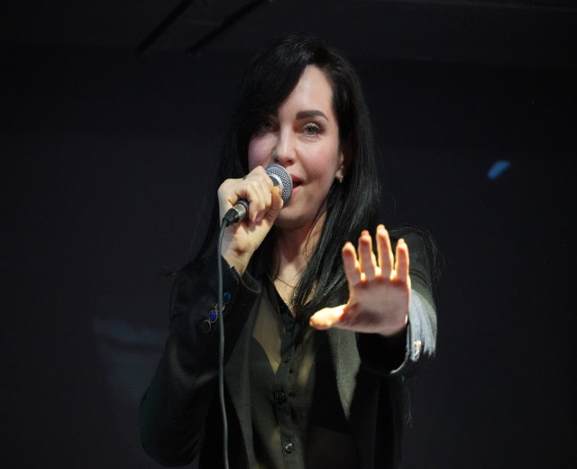 Eine Frau in einem schwarzen Kleid singt leidenschaftlich in ein Mikrofon auf einer Bühne, ihre Augen geschlossen und das Haar zurückgebunden, beleuchtet von einem hellen Licht.