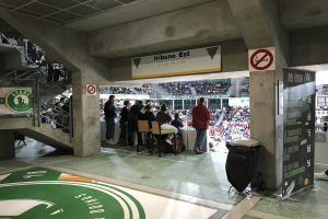 Gruppe von Menschen, die vor einer sitzenden Menge in einem Stadion mit Schildern stehen, die "Boston Celtics vs New York Jets" lesen, ein Müllcontainer, ein Textbrett, eine Treppe mit Geländern und eine Decke mit Deckenleuchten.