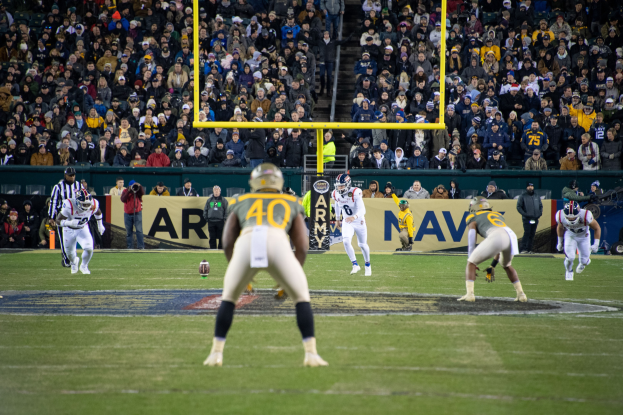 Fussballspieler in Sportkleidung und Helmen konkurrieren auf einem Feld mit Zuschauern im Hintergrund, Stadionmasten und Textanzeigetafeln.