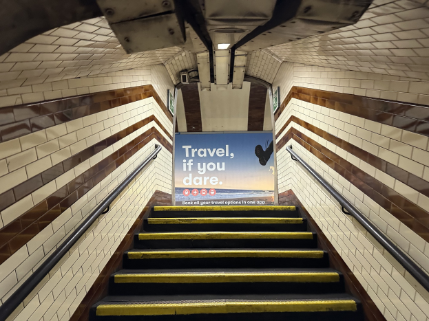 Eine Treppe in einer U-Bahn-Station mit einem "Reise, wenn du wagst"-Schild oben, flankiert von Geländern und sichtbaren Stationswänden.