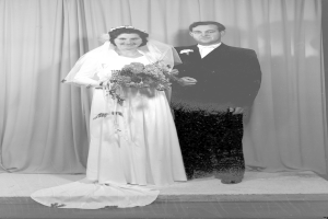 Ein Schwarz-Weiß-Foto eines Brautpaares auf einer Bühne, wobei die Braut einen Blumenstrauß hält und der Bräutigam einen Anzug trägt, mit einem Vorhang dahinter.