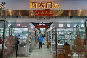 Innenansicht einer chinesischen Apotheke mit Glaswänden, Regalen voller Artikel, Kartons auf dem Boden, einer Pflanze links und einer Namensschild oben.