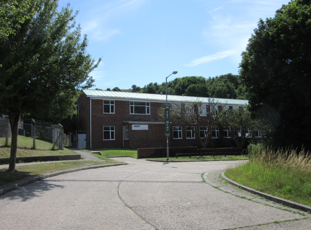 Ein Schulgebäude mit Fenstern, einem Straßenpfahl, einem Schild und einer Gruppe von Bäumen, umgeben von saftigem Gras und einem Zaun, unter einem bewölkten Himmel.