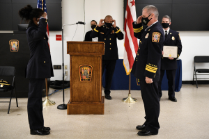 Eine Gruppe uniformierter Personen mit Masken auf einem Podium während einer Amtseinführung für einen neuen Feuerwehrchef, der eine Urkunde hält, mit Flaggen, Stühlen, Tischen und einer Wand im Hintergrund.