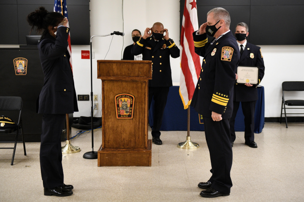 Eine Gruppe uniformierter Personen mit Masken auf einem Podium während einer Amtseinführung für einen neuen Feuerwehrchef, der eine Urkunde hält, mit Flaggen, Stühlen, Tischen und einer Wand im Hintergrund.