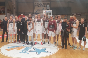 Mannergruppe mit Kindern auf einem Basketballfeld mit Schildern "Hala Osir Starga", sitzende Zuschauer, ein Basketballkorb und Deckenbeleuchtung.