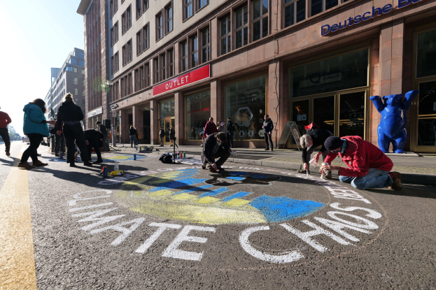 Menschen sitzen vor einem Gebäude mit Fenstern und Namensschildern auf dem Boden, umgeben von Flaschen und anderen Gegenständen, während sie an einer Klimawandel-Demo in Berlin teilnehmen, mit Bäumen und einem klaren blauen Himmel im Hintergrund.