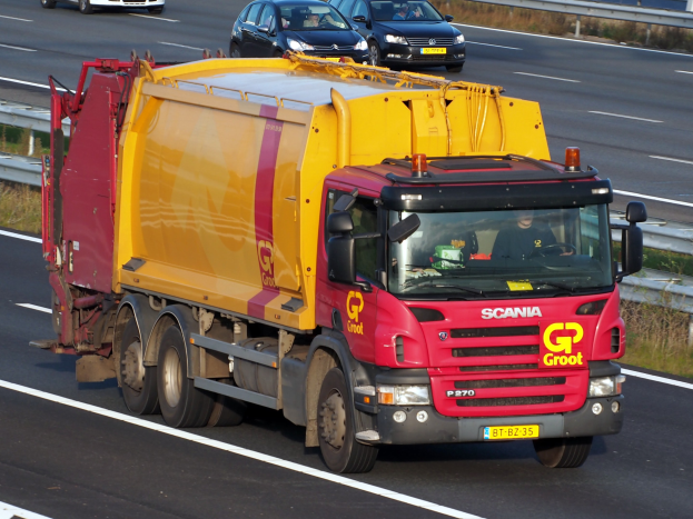 Ein gelber und roter Müllwagen auf einer Autobahn neben anderen Fahrzeugen, mit Gras und einem Geländer am Straßenrand.