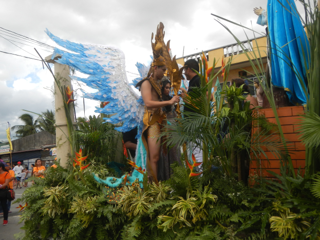 Eine Gruppe von Menschen geht neben einem mit Pflanzen und Blumen geschmückten Festwagen die Straße entlang, mit kostümierten Personen auf dem Wagen, Gebäuden, Bäumen, Strommasten mit Drähten und einem bewölkten Himmel im Hintergrund.