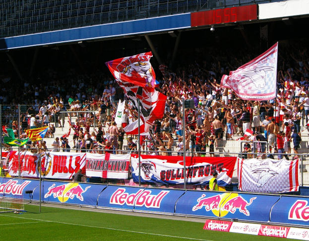 Ein Fußballfeld mit einem Tor auf der linken Seite, einer Zuschauertribüne, Bannern, Pfosten und Fahnen im Vordergrund und einem Gebäude im Hintergrund.