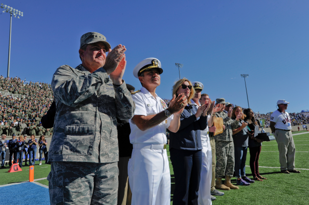 Eine Gruppe von Menschen auf einem Fußballfeld klatschen, mit Zuschauern auf den Rängen, Lichtmasten und einem klaren blauen Himmel im Hintergrund.