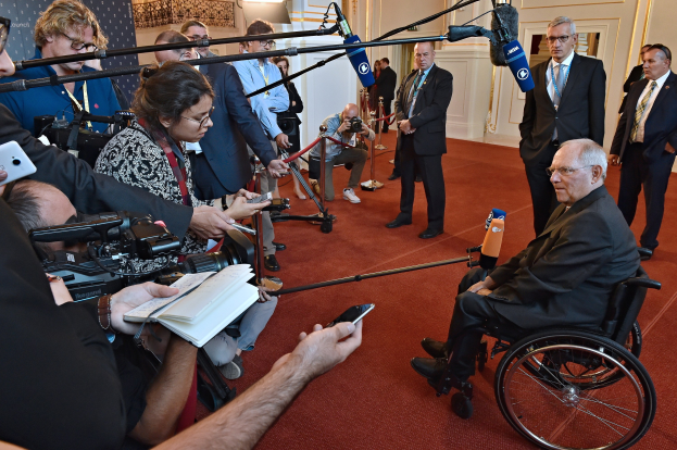 Ein Mann im Rollstuhl spricht zu einer Gruppe von Reportern mit Kameras, Handys und Mikrofonen mit Ständern, einem Banner, Scheinwerfern und einer Wand im Hintergrund.