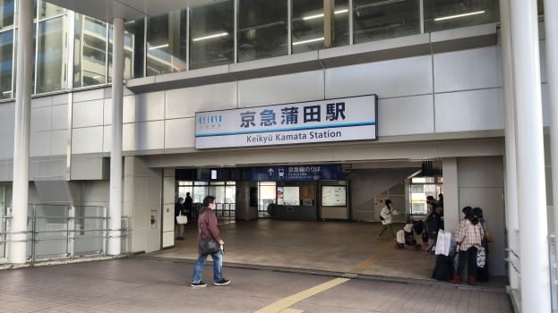 Menschen mit Taschen durch eine Bahnhofshalle gehen, ein Schild mit Text, Säulen, Geländer, Glaswände und Deckenlampen; ein Mann in der Mitte mit Schuhen.