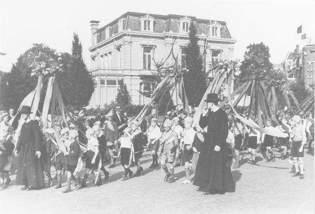 Schwarzes und weißes Bild einer Gruppe von Menschen, die eine Straße entlanggehen, einige halten Fahnen, mit Hüten, Bäumen, Gebäuden und einem Maibaum im Hintergrund.