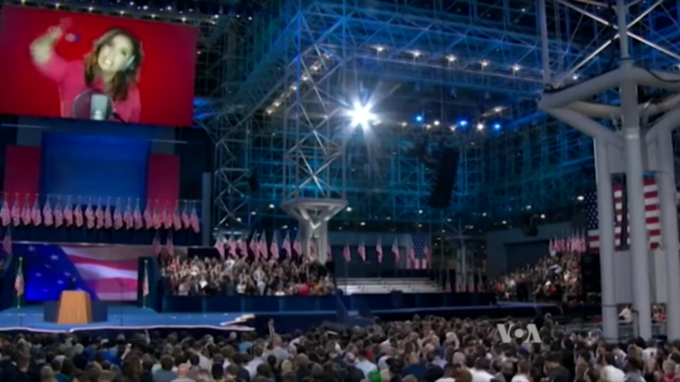 Eine große Menschenmenge sitzt vor einer Bühne mit einem Rednerpult, Mikrofon, Fahnen, einem Bildschirm mit einer Frau und Lichtausrüstung und wartet auf eine Präsidentschaftsdiskussion.