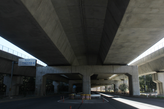 Autobahn unter einer Brücke mit Absperrkegeln, Barrieren und textbeschrifteten Tafeln, gesäumt von Geländern, Bäumen und einem klaren blauen Himmel im Hintergrund.