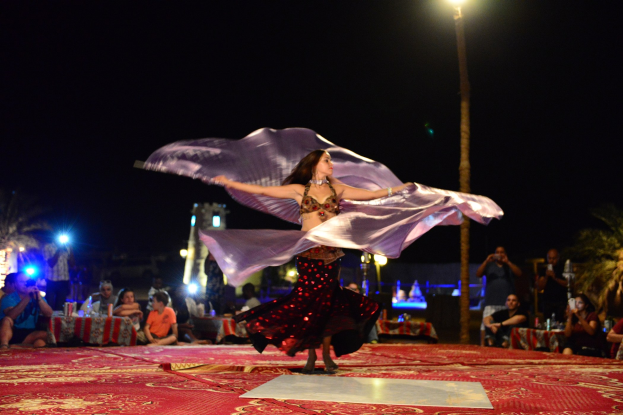 Eine Frau in einem Bauchtanzkostüm tanzt auf einem roten Teppich, umgeben von einer Menge Fotografen und Zuschauern, mit Bäumen, Lichtern, einem Pfahl und Himmel im Hintergrund.