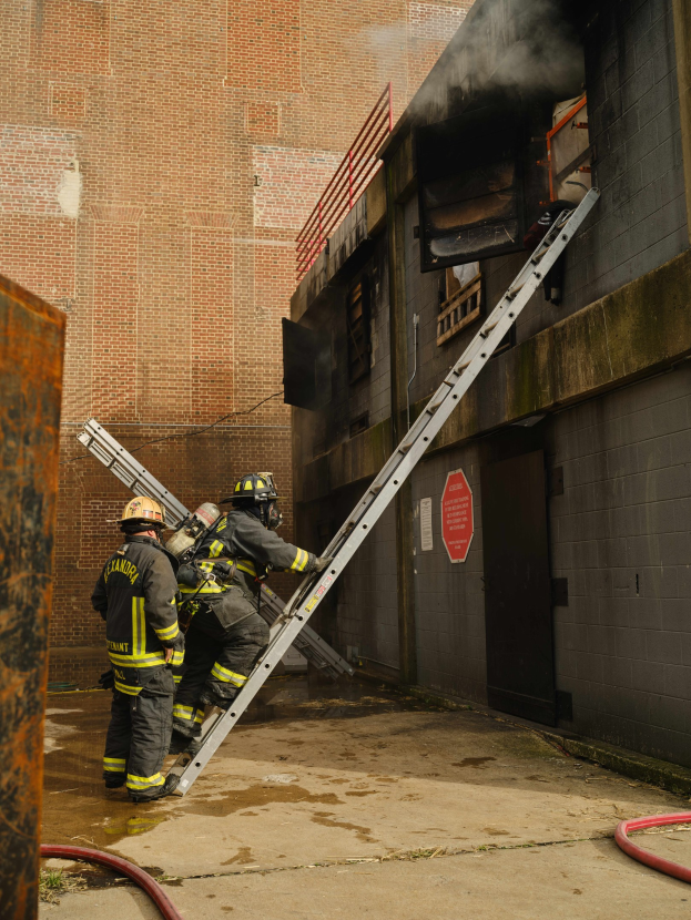 Feuerwehrleute mit Helmen klettern eine Leiter zu einem rauchgefüllten Gebäude hoch, mit einem Metallobjekt links und Rohren am Boden.