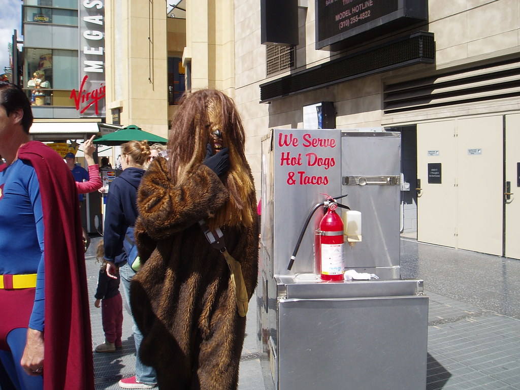Eine Menschenmenge, darunter einige in Kostümen, versammelte sich vor Gebäuden mit sichtbarer Beschriftung in der Nähe einer Box mit Schrift und einem Feuerlöscher.