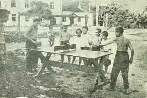 Eine Gruppe von Kindern spielt Tischtennis vor einem Gebäude mit Fenstern, umgeben von Bäumen und Pfählen, in einem Schwarz-Weiß-Bild mit Text unten.