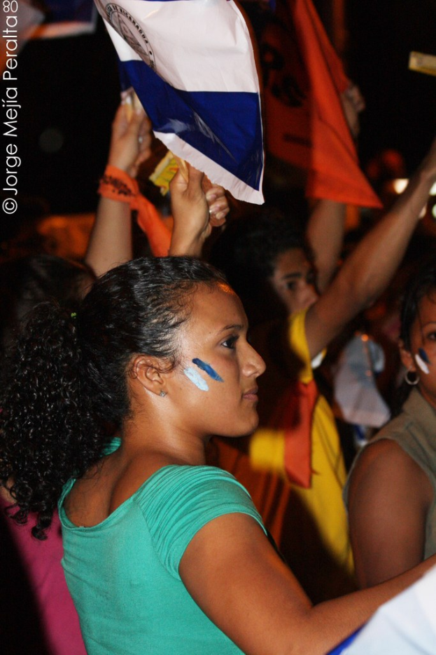 Frauen in grünen T-Shirts, die bei einer Protestaktion stehen und weiße Schilder halten, mit vielen anderen Mädchen im Hintergrund.