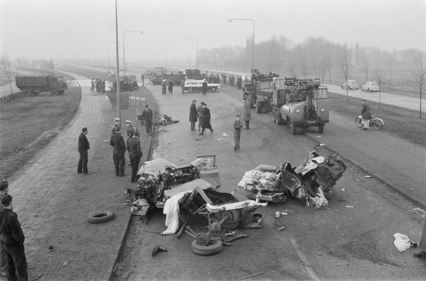 Schwarze und weiße Szene eines Autounfalls am Straßenrand mit mehreren Fahrzeugen, darunter ein Landfahrzeug, und einer Gruppe von Menschen, die darum herumstehen, mit Laternenpfählen, Bäumen und dem Himmel im Hintergrund.
