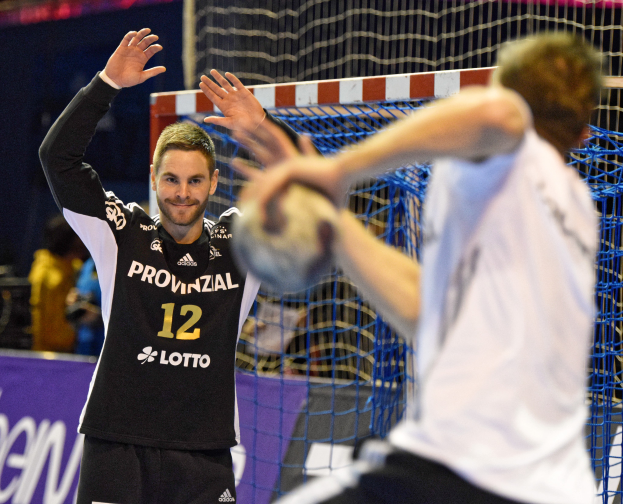 Zwei Männer spielen Handball vor einem Tor mit Netz und einer Fahne mit Text im Hintergrund und einigen Zuschauern in einer Stadion.